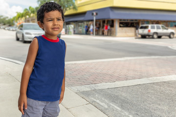 A young boy stands alone on the street corner with a concerned look. He's focused on looking down the street while a car approaches from behind.