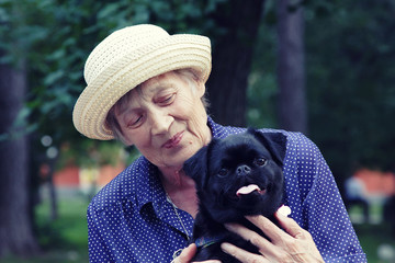 Portrait of elderly woman holds a small dog in her arms and talks to dog. The dog is showing emotions. The breed of dog is a pug.