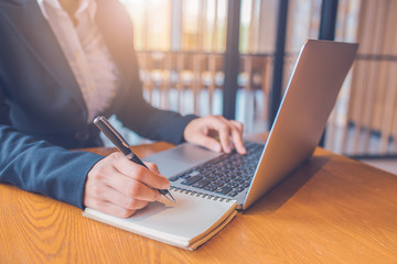 Business women are taking notes on paper with a black pen, and she is using a laptop computer on a...