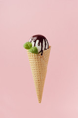 White creamy ice cream in crisp waffle cone with chocolate sauce, fresh green mint leaf on pastel pink background.