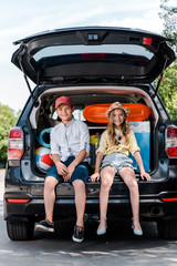 happy kid in cap sitting near sister in straw hat and looking at camera