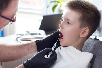 Dentysta w czarnych gumowych rękawiczkach trzyma w dłoni strzykawkę z igłą i robi zastrzyk chłopcu siedzącemu na fotelu dentystycznym z otwartymi ustami. - obrazy, fototapety, plakaty