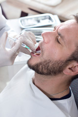 Lekarz stomatolog robi zastrzyk ze znieczuleniem w dziąsło pacjenta. Twarz mężczyzny z otwartymi ustami na fotelu stomatologicznym - obrazy, fototapety, plakaty