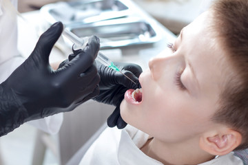 Dentysta w czarnych gumowych rękawiczkach trzyma w dłoni strzykawkę z igłą i robi zastrzyk chłopcu siedzącemu na fotelu dentystycznym z otwartymi ustami. - obrazy, fototapety, plakaty