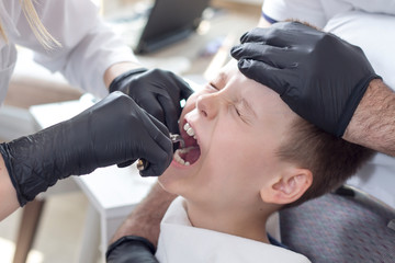 Twarz dziecka z grymasem bólu. Lekarz wyrywa zęba a pielęgniarka trzyma głowę dziecka przyciśniętą do zagłówka fotela dentystycznego.