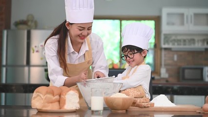 Cute little Asian boy and beautiful mother sifting dough flour with sifter sieve colander in home kitchen on table for prepare to baking bakery and cake. Thai kids playing with flour as chef funny