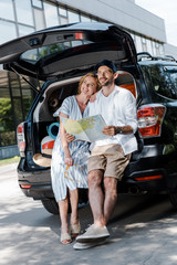 cheerful man standing near car and beautiful woman while holding map