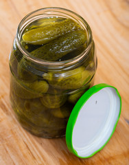 Opened glass jar with pickled cucumbers on wooden background