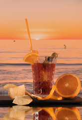 Exotic cocktail with olives, orange slices and French fries with the waves of the sea at the background - On a glass table