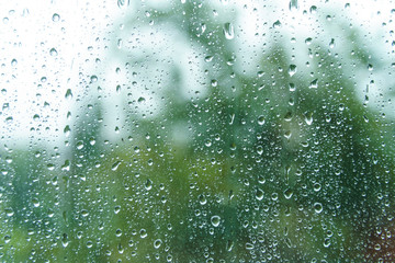 Rain drops on rainy day on outside window glass with blurred edges. Rain outside window pane in spring day. Water drops flow down the surface. Droplets on window with green tree in background.