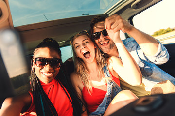 Three best friends enjoying traveling in the car, sitting in rear seat and having lots of fun making selfie on a road trip.