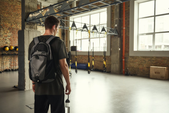 Morning workout. Back view of young man with backpack standing in empty gym
