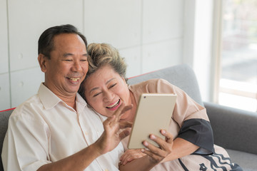 Senior happy Asian couple sitting on couch and do activity together using smart tablet for entertainment