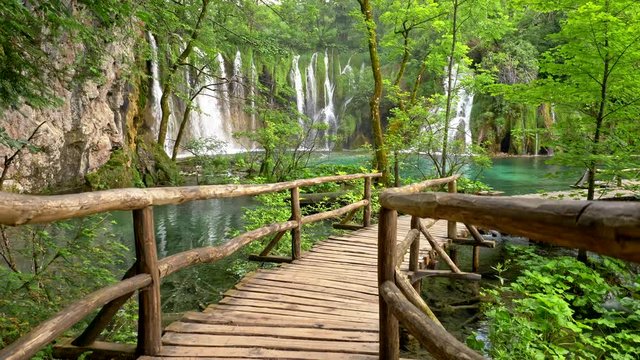 Boardwalk in Plitvice Lakes National Park, Croatia. Transparent turquoise colored waters of the lake are having ripples. Many small waterfalls flowing into the lake. All this surrounded by rich green