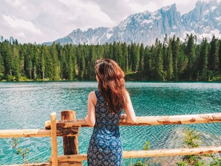 A brunette girl with long hair in a long dress stands near the mountain lake Carezza, a romantic beautiful place, azure pure water. Mountain summer landscape in the Dolomites in Northern Italy. 