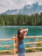A brunette girl with long hair in a long dress stands near the mountain lake Carezza, a romantic beautiful place, azure pure water. Mountain summer landscape in the Dolomites in Northern Italy. 