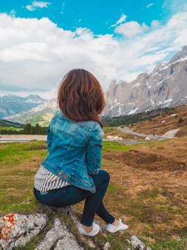 Brunette girl with long hair in a shirt sitting on a big boulder on the background of Dolomite mountains. Mountain summer landscape in the Dolomites in Northern Italy. 