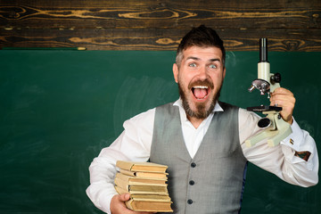 Scientifics research. Male teacher of biology holds books and microscope. Student in classroom. Biology or chemistry lesson. Scientist holds books and microscope. Chalkboard on background, copy space.
