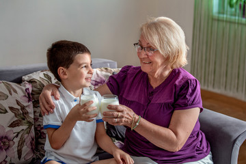 Obraz na płótnie Canvas Happy little grandson drinks milk with straw in glass with grandmother. Use of calcium. Beautiful grandma with grandson drinking milk, healthcare concept.