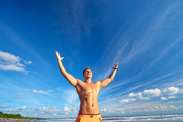Fototapeta premium Victory and freedom. Young handsome strong man raising hands up on the beach against sky.