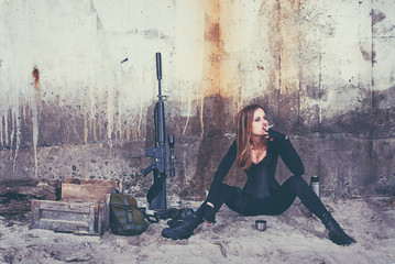 A badass lady in a black catsuit smokes a cigarette while sitting near her combat equipment.