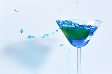 Cocktail with blue liquid in glass. Glass with blue water pouring with liquid with splashes and drops. Martini glass filling with alcohol with splashes on white background. Refreshing drink concept.