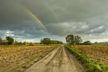 A dirt road through fields and two rainbows in the dark sky