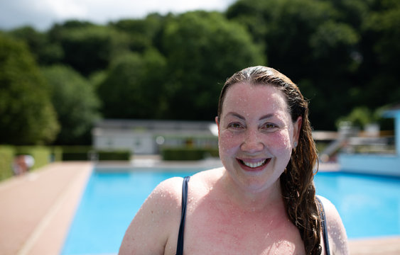 close up portrait of a confident large plus size woman in front of public swimming pool smiling looking at camera with wet hair on a sunny summer day