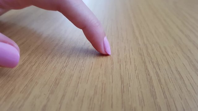 Female finger with white skin and pink short nail scratching a wooden table, close-up
