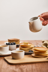 cropped view of man holding jug with syrup upon plate with pancakes and blueberries near cup of coffee and bowl with honey isolated on grey