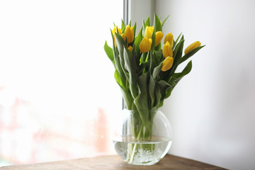 A bouquet of yellow tulips in a vase on the windowsill. A gift to a woman's day from yellow tulip flowers. Beautiful yellow flowers in a vase by window.