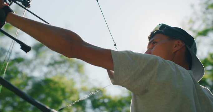 Slow motion close up of young man with professional equipment is practising archery with a bow in a woods in a sunny day. 