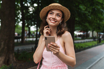 Photo closeup of glamorous young woman using cellphone and earphones while walking through green park