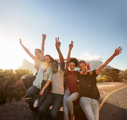 Five millennial friends on a road trip taking a break, leaning on the car waving to camera