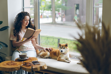 Female student is reading book and stroking pet dog sitting on window sill in cafe enjoying hobby...