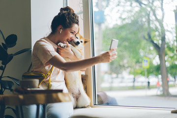 Cheerful lady taking selfie with doggy in cafe using smartphone camera sitting on window sill...