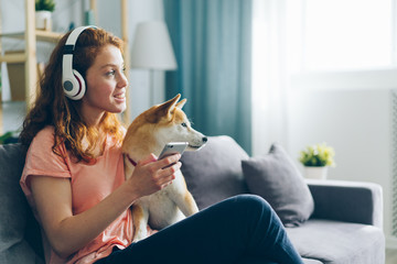 Beautiful woman in headphones is listening to music and petting pedigree dog sitting on couch...