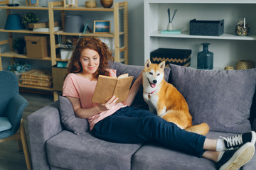 Pretty young woman with long curly red hair is reading book and caressing shiba inu dog sitting on couch at home. Lifestyle, youth culture and animals concept.
