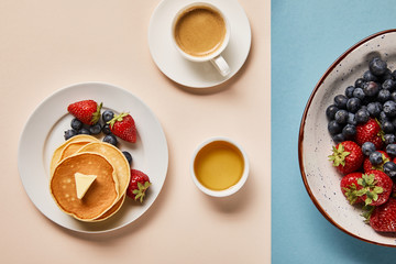 top view of pancakes with berries on plate, bowl with honey, cup of coffee and plate with berries on pink and blue background