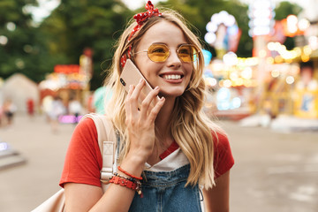 Smiling pleased young cheery blonde woman in amusement park talking by mobile phone.