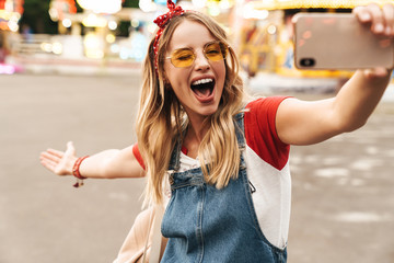 Image of joyful blonde woman taking selfie photo on cellphone in front of colorful carousel at amusement park