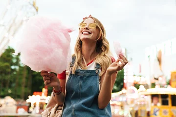 Photo sur Plexiglas Parc dattractions Image of joyful blonde woman eating sweet cotton candy while walking in amusement park