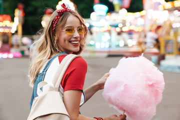 Image of smiling young woman eating sweet cotton candy while walking in amusement park