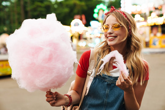 Image of happy blonde woman eating sweet cotton candy while walking in amusement park