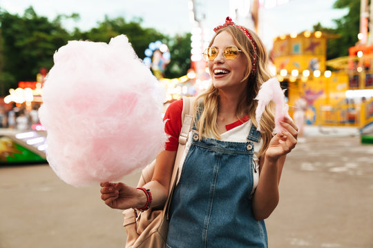 Image of smiling blonde woman eating sweet cotton candy while walking in amusement park