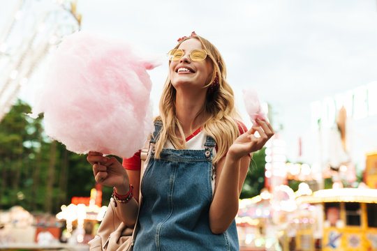 Image of joyful blonde woman eating sweet cotton candy while walking in amusement park