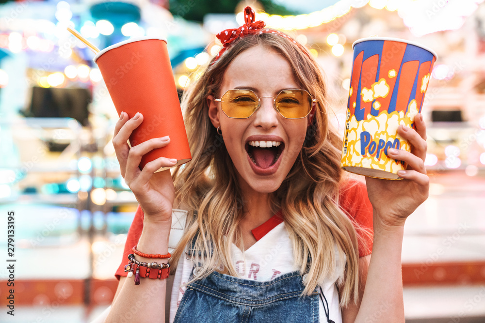 Wall mural Image of joyful charming woman holding popcorn and soda paper cup while walking in amusement park