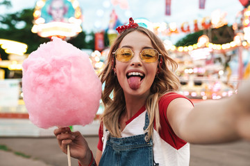 Afbeelding van een positieve blonde vrouw die zoete suikerspin vasthoudt terwijl ze een selfie-foto maakt in het pretpark