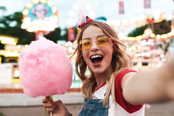 Zelfklevend Fotobehang Amusementspark Afbeelding van een opgewonden blonde vrouw die zoete suikerspin vasthoudt terwijl ze een selfie-foto maakt in het pretpark