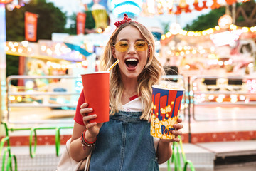 Fototapeta Image of excited charming woman holding popcorn and soda paper cup while walking in amusement park obraz